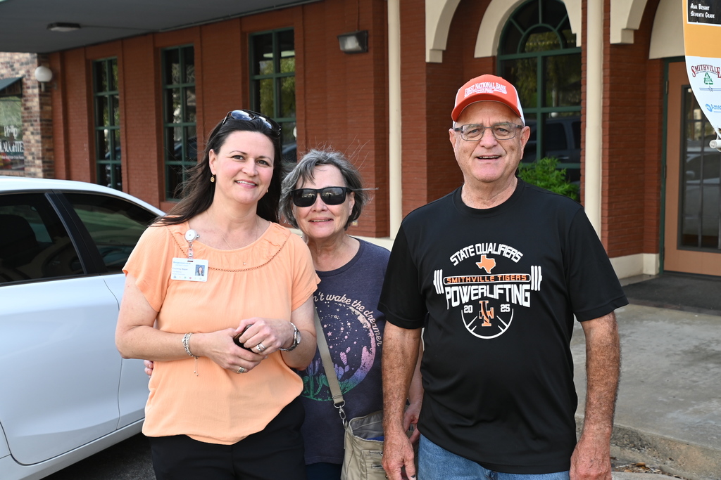 Three people stand outside, one in orange, one in black, and one in blue. A car and a building are behind them.