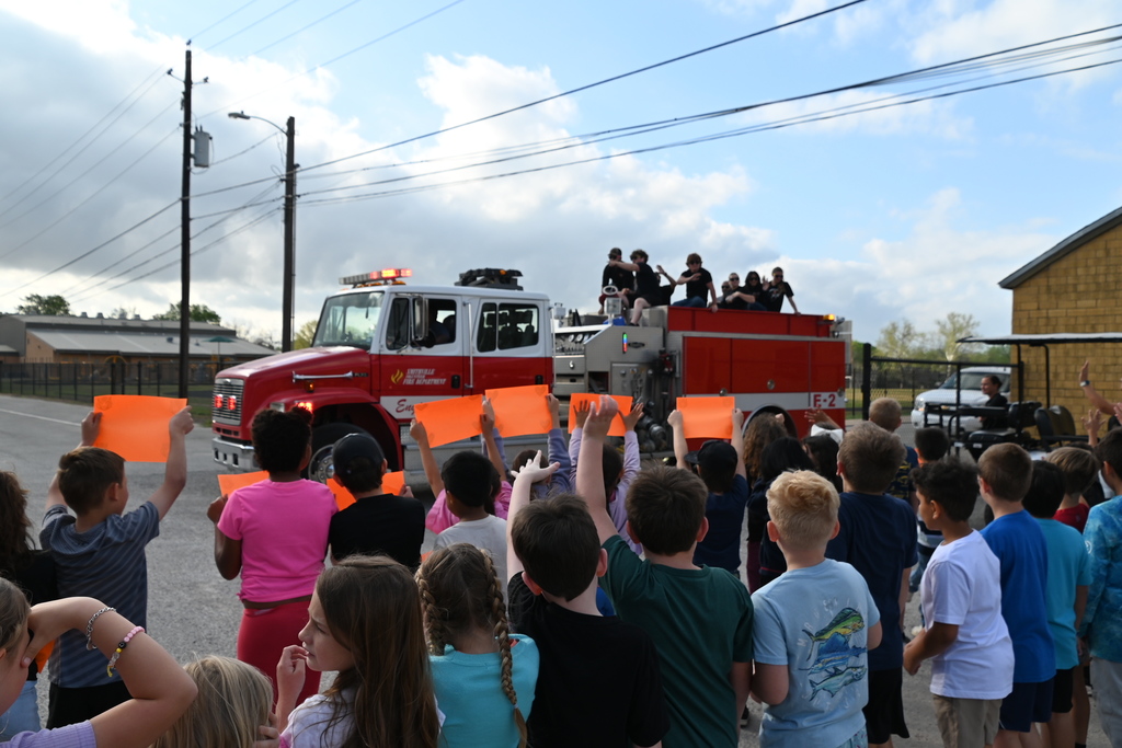A group of children hold up orange signs in front of a red fire truck. The truck is parked on a street.