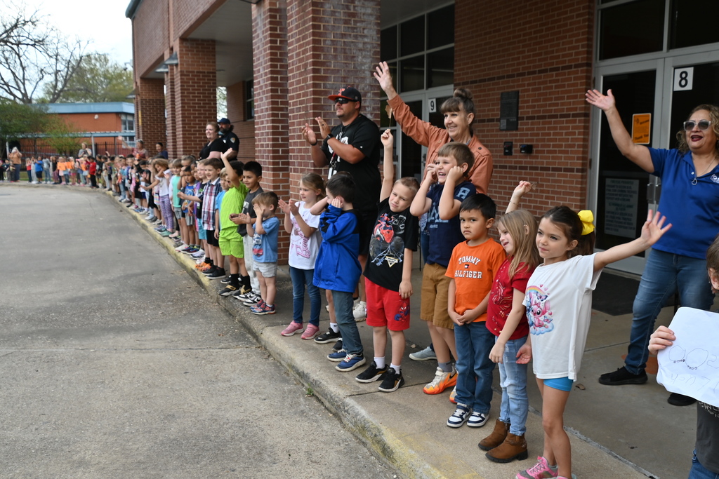 Group of children and adults standing outside a building, waving and smiling, on a sunny day.