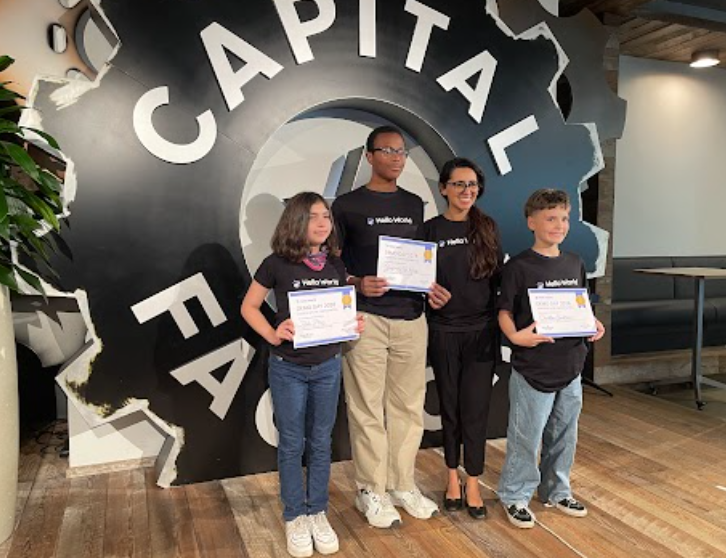 Four people stand behind a sign with a broken gear and text reading "CAPITAL FACTORY" holding certificates