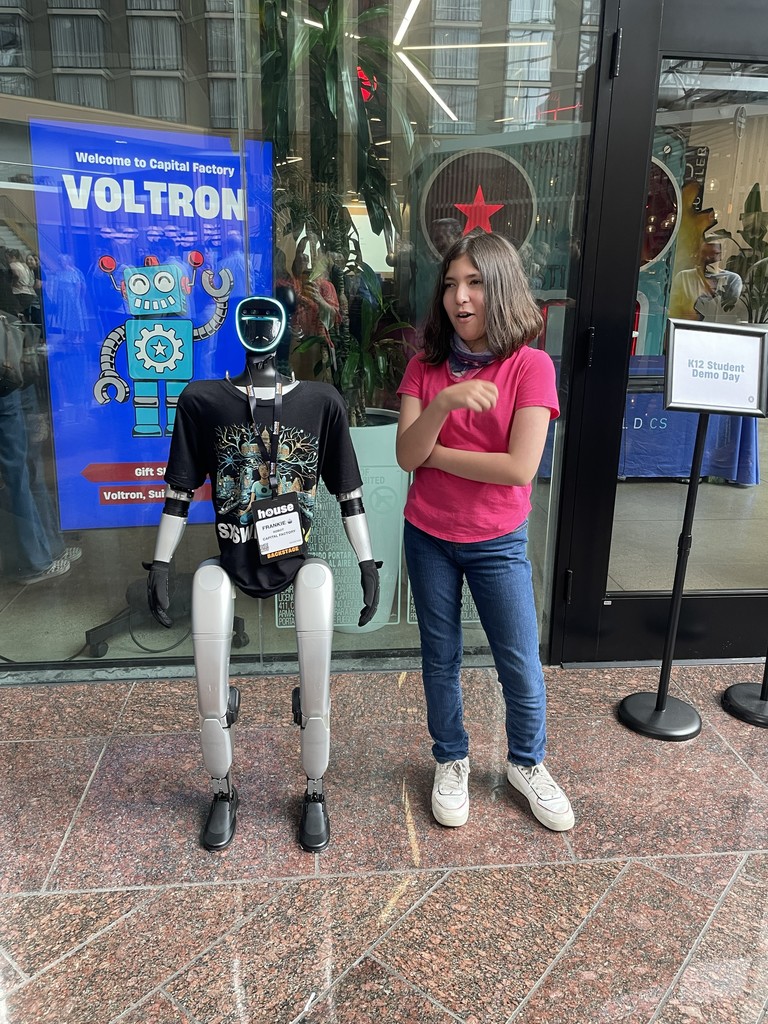 A girl in a pink shirt and jeans stands beside a white robot in front of a building with a sign that says "VOLTRON".