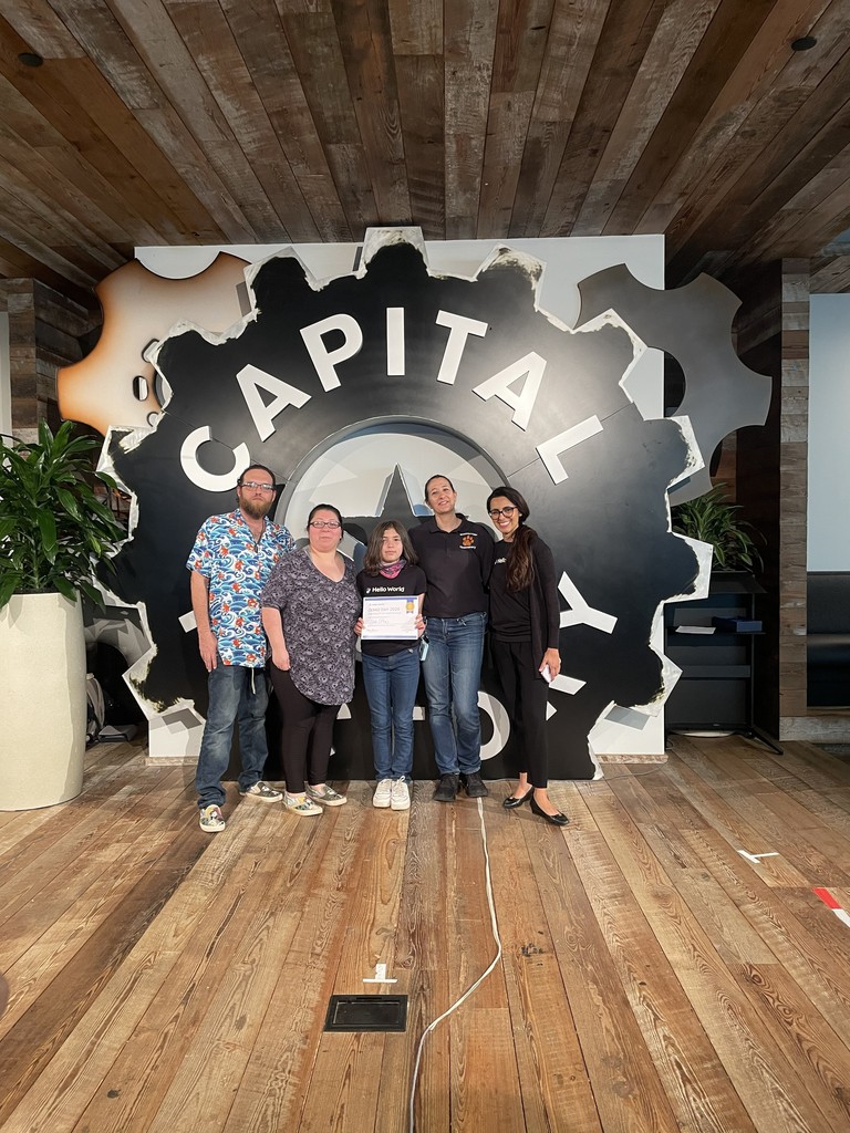 Five people stand in a room with a wooden floor and a large sign reading "CAPITAL" in the background.