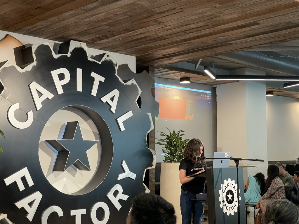 A woman speaks at a podium in front of a sign reading "CAPITAL FACTORY" with a star and gear design.