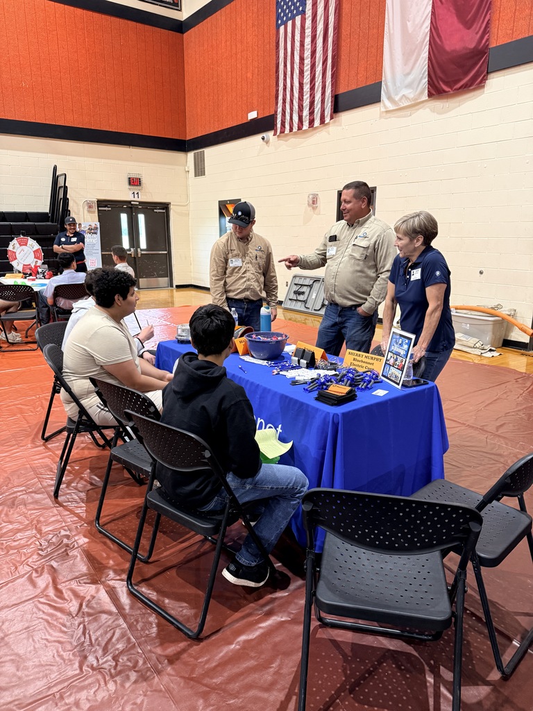 People sit around a table with food. A man stands and speaks to them. Flags hang on a wall.