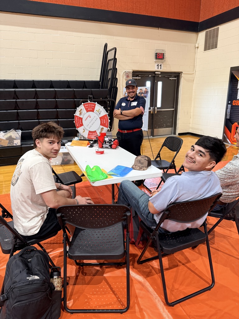Two young men sit on chairs with a table in a gym. A man stands behind them.