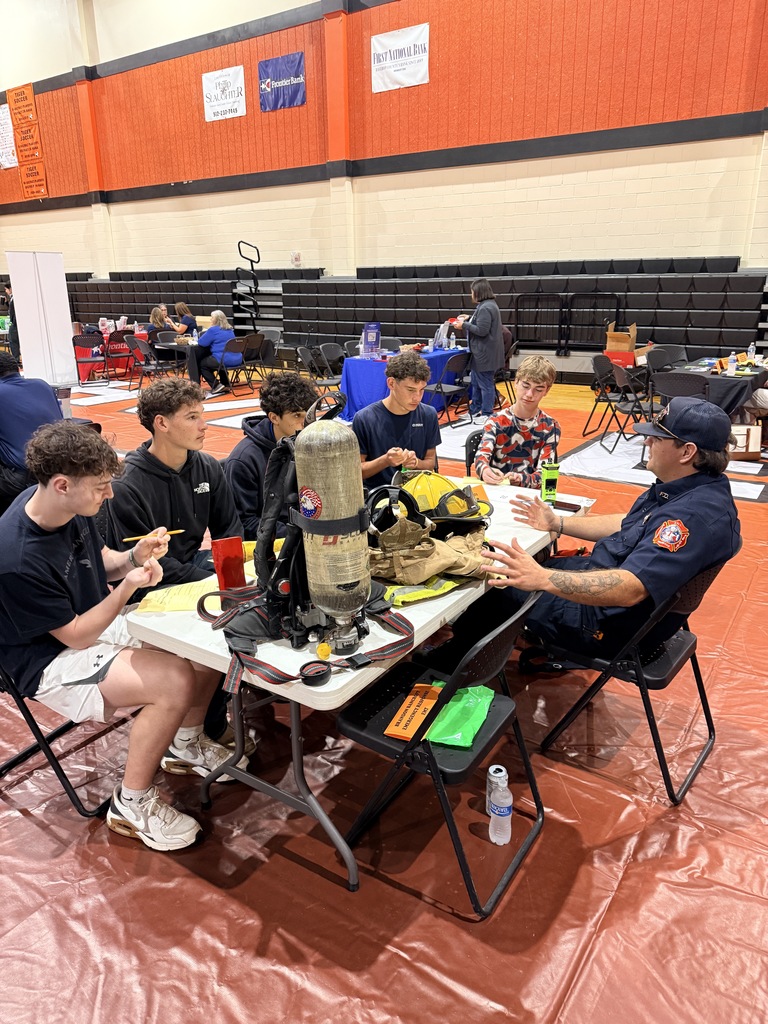 People in blue shirts sit around a table with equipment inside a gym with red floor covering.