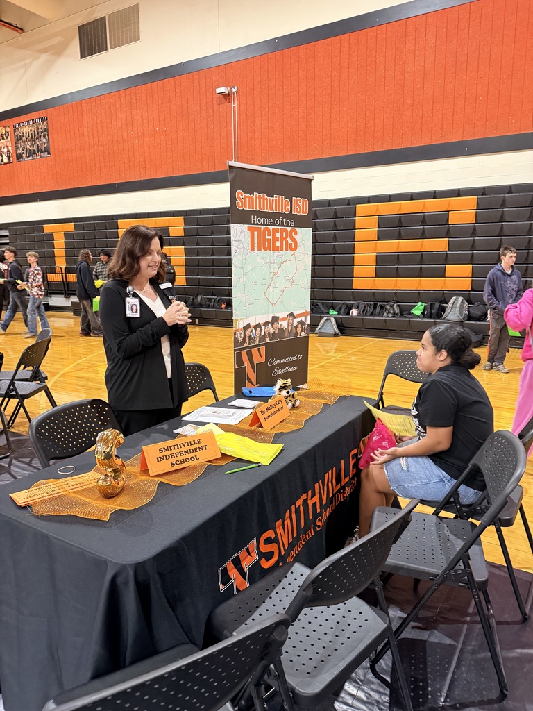 Two people at a table in a gymnasium with a sign for AP Smithville in front of them.