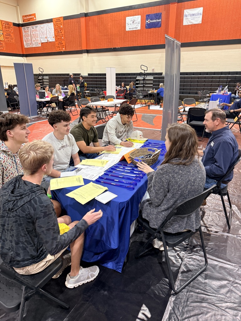 A group of students sits around a table with papers and a man at a career fair.