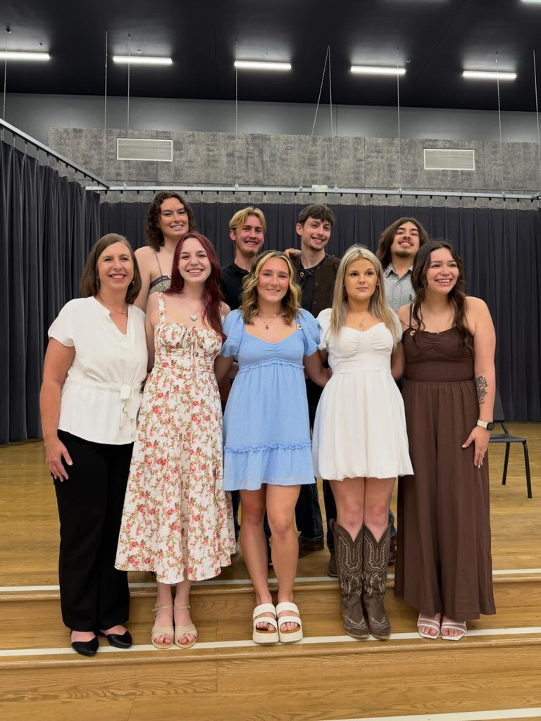 A group of people in various dresses and shoes pose together on a stage with a black curtain background.