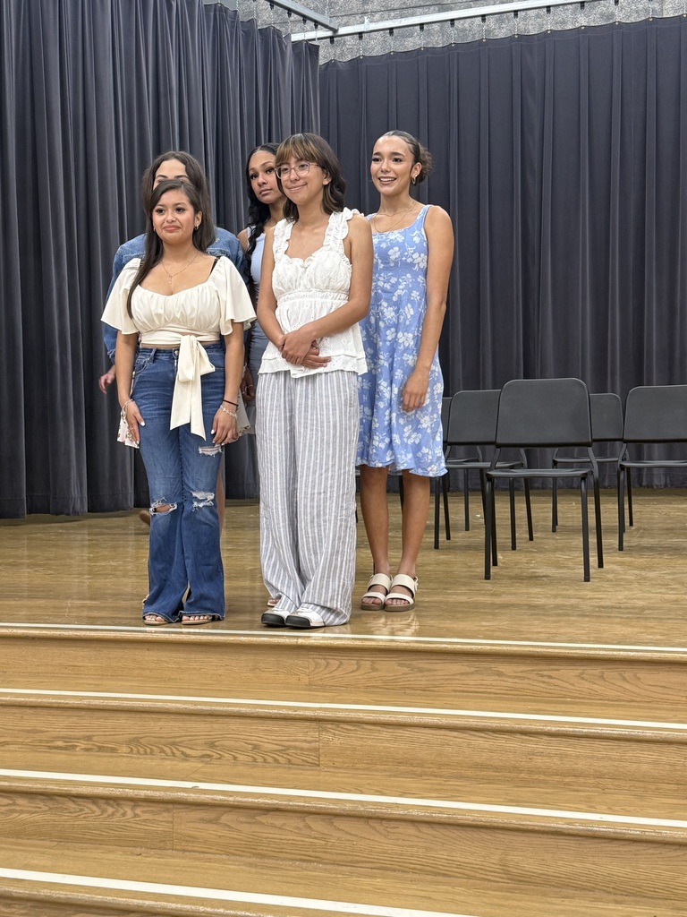 Four women stand on stage with wooden steps and black curtains in the background. Empty black chairs line the back.