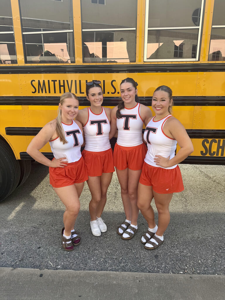 four cheerleaders stand in front of a bus in their cheer uniforms