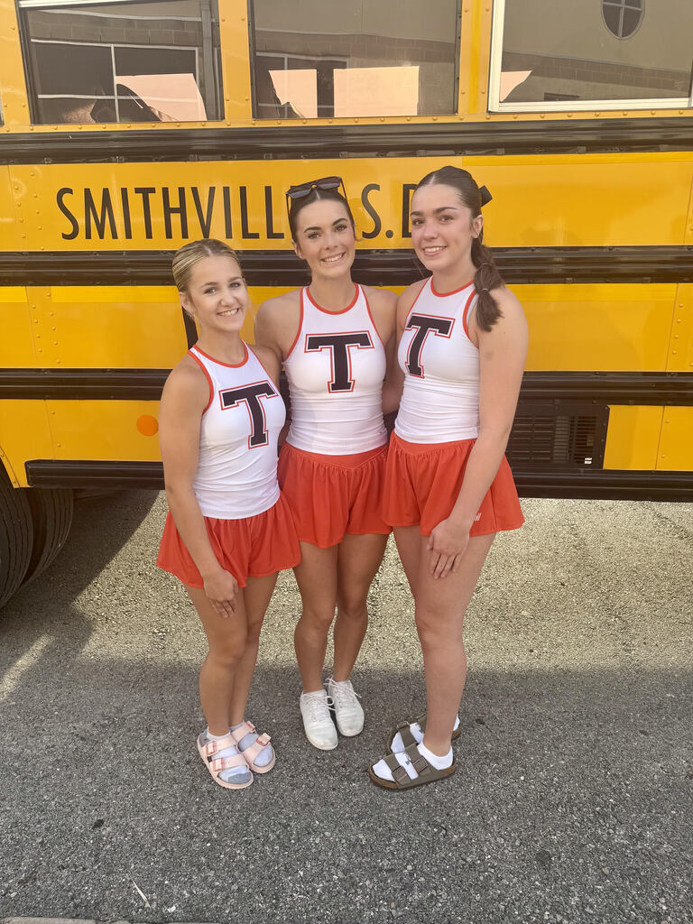 three cheerleaders stand in front of a bus in their workout outfits