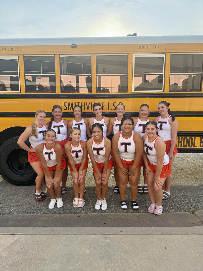 Eleven cheerleaders pose in front of a yellow school bus, all wearing matching white and red uniforms.