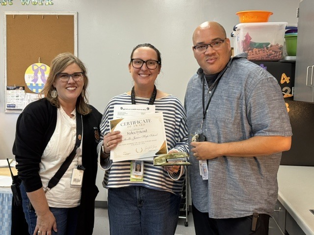 Three people in a kitchen. One holds a certificate, while two others smile. A bulletin board and fridge are in the background.