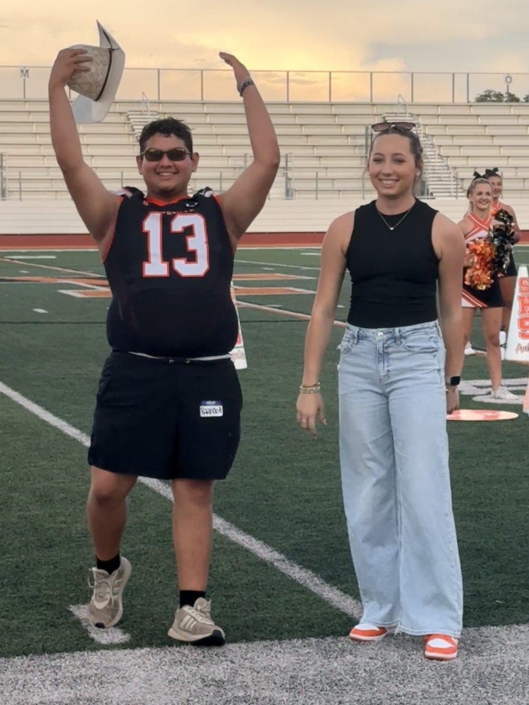 Two people stand on a football field. A man in a jersey with the number 13 and a woman in a sleeveless top and jeans.