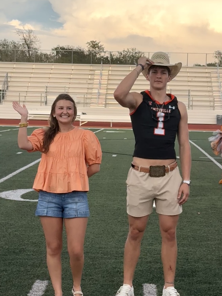 A woman and man stand on a football field. The woman wears an orange top and denim shorts. The man wears a cowboy hat and a black jersey.