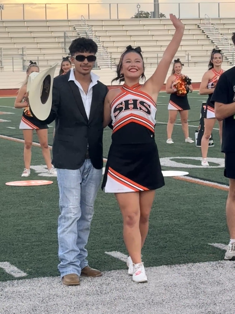 A man in a suit and a woman in a cheerleading uniform stand on a field, smiling. Behind them, several cheerleaders in uniforms are visible.