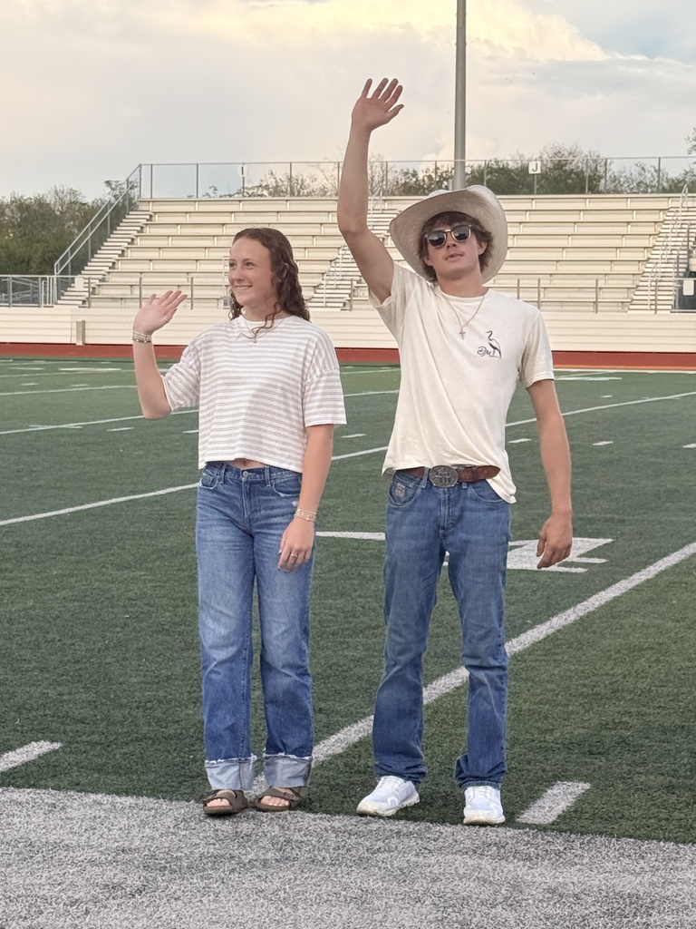 Two people on a field with bleachers in the distance. Both are dressed in casual attire.