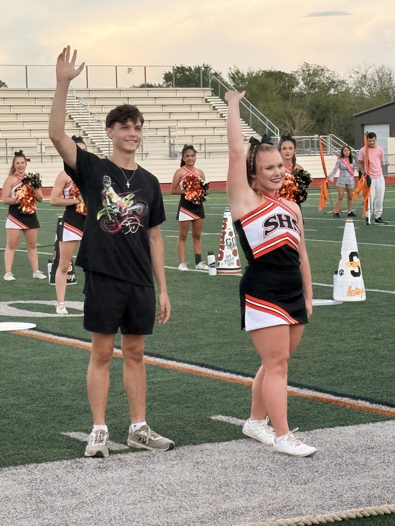 A man and woman stand waving on a football field. Cheerleaders are in the background.