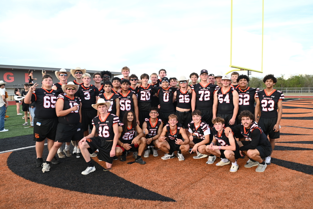 Football team wearing black jerseys with numbers 16, 56, 20, 26, 72, 64, 62, 23, and 65, posing on an athletic field.