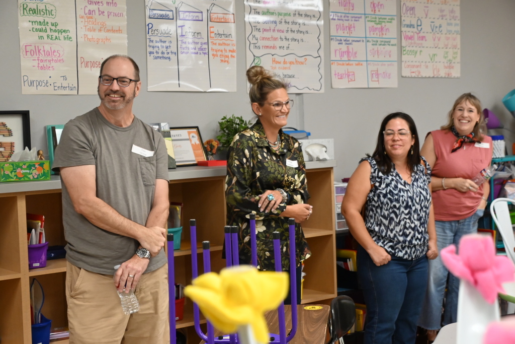 Four people stand in a room with a bookshelf and posters on the wall. One holds a colorful object.