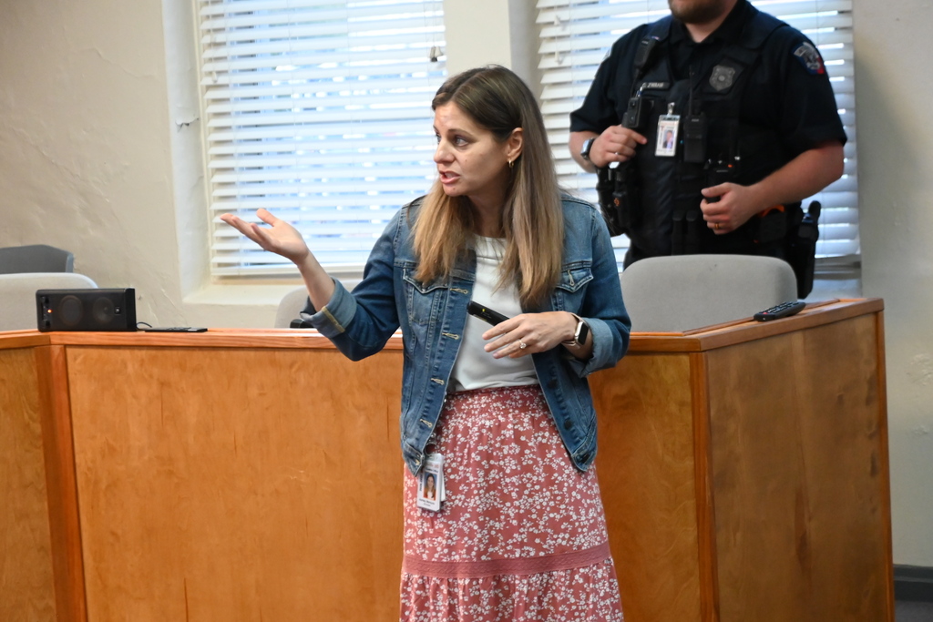 A woman in a denim jacket and pink skirt stands in front of a wooden desk. A person in uniform stands behind her.