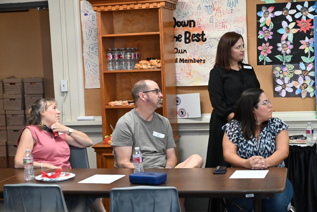 Four people seated at a table. One woman stands behind the table. A woman with glasses speaks.