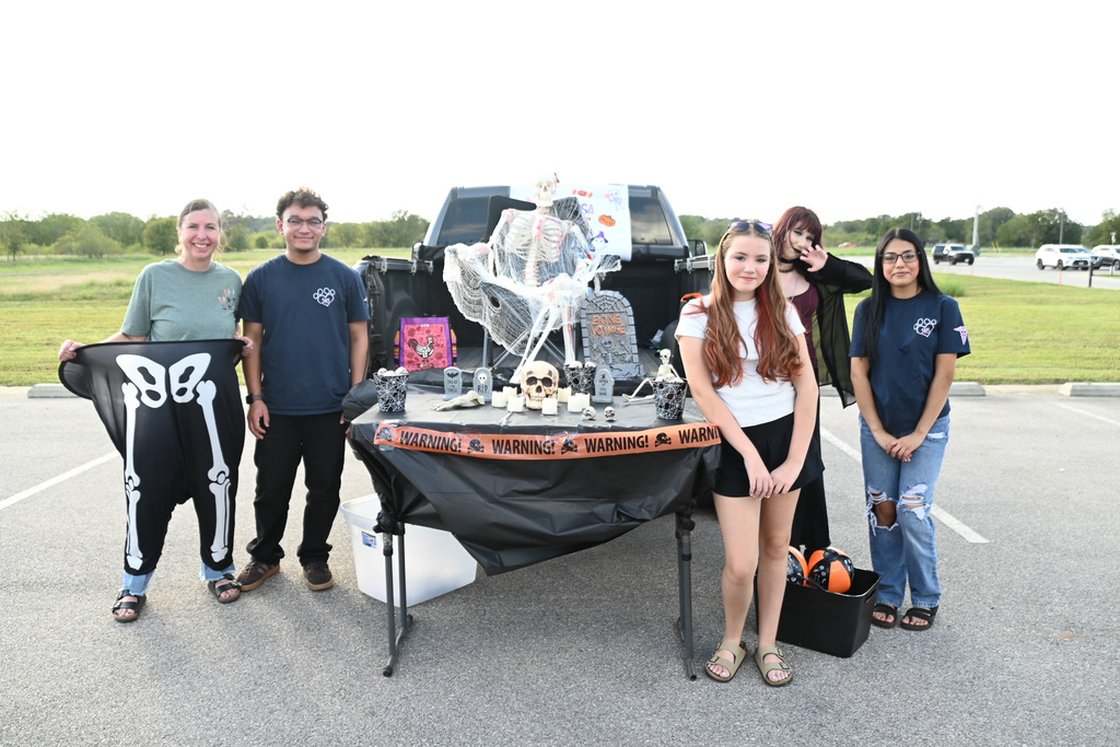 Group of six people in outdoor parking lot. Four adults and two children standing behind table with Halloween decorations.