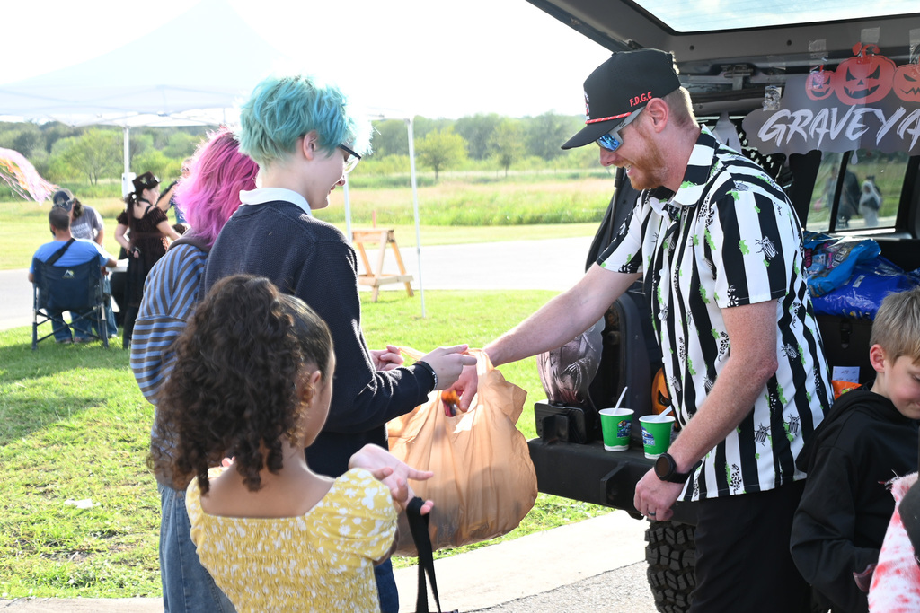 A man in a striped shirt hands out bags to children at a Halloween trunk-or-treat.