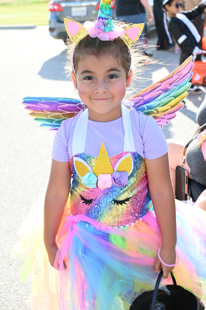 Young girl in colorful unicorn costume with rainbow wings and tutu, standing in a parking lot.