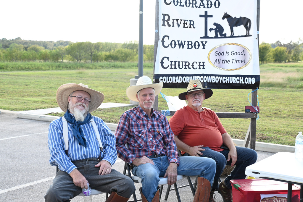 Three men in cowboy hats and boots sit on chairs outside, with a sign for Colorado River Cowboy Church in the background.