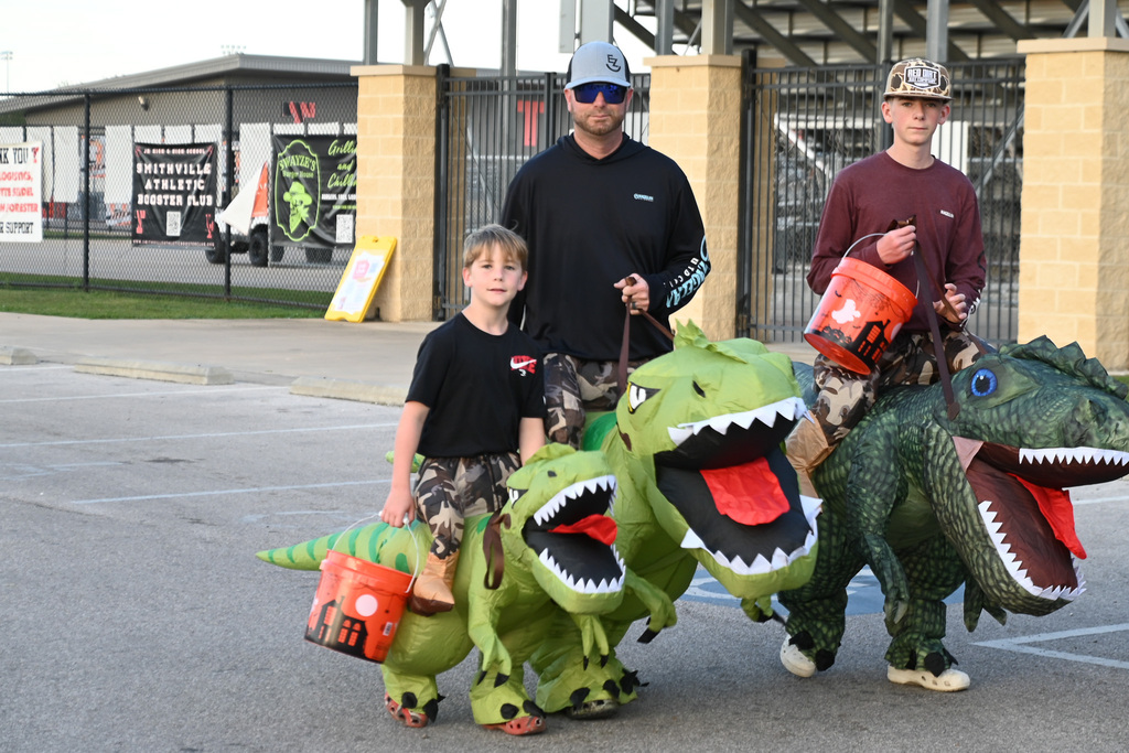 Four people dressed in large dinosaur costumes holding orange buckets pose in a parking lot.