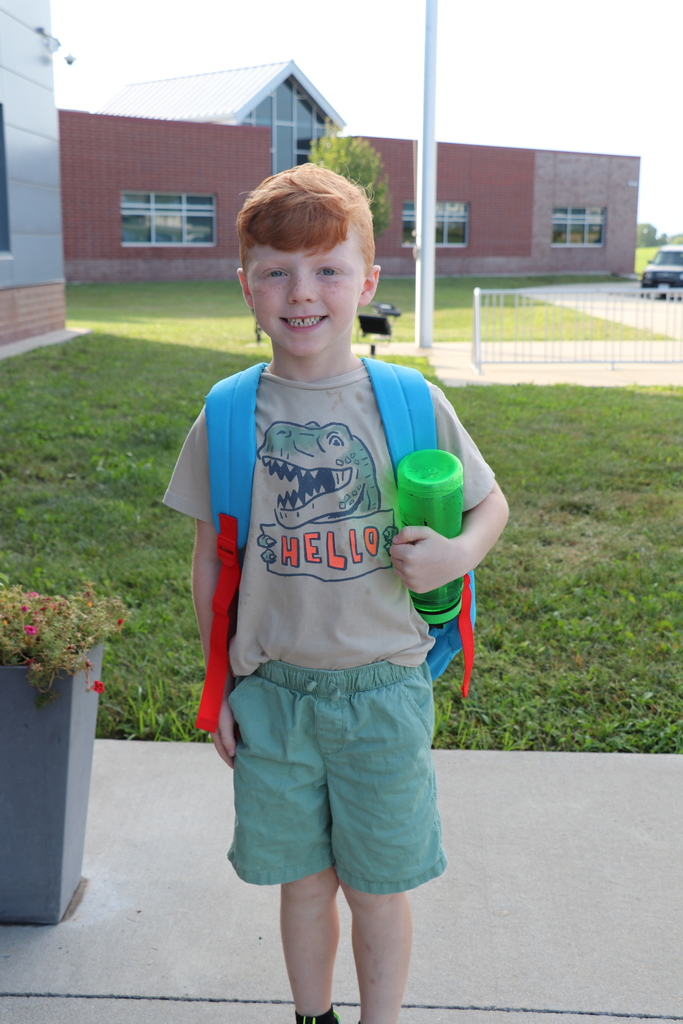 A boy poses in front of the school with his backpack on and water bottle in his hand