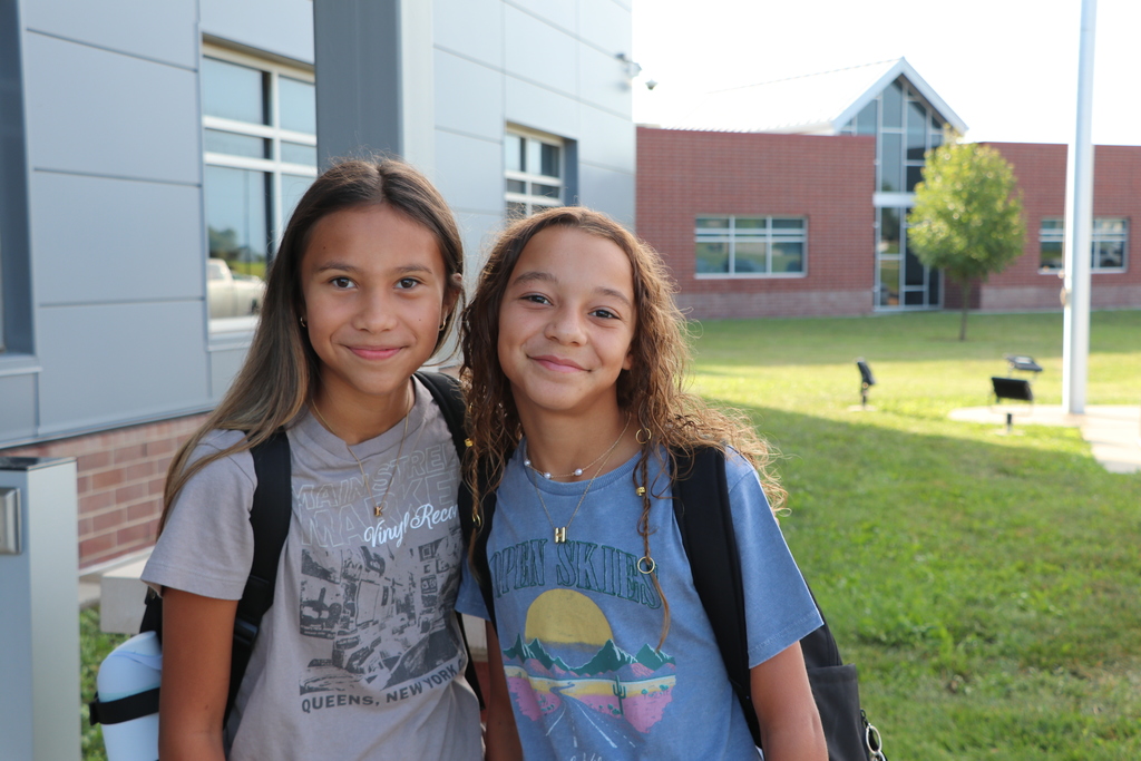 Two girls pose in front of the school with their backpacks on.