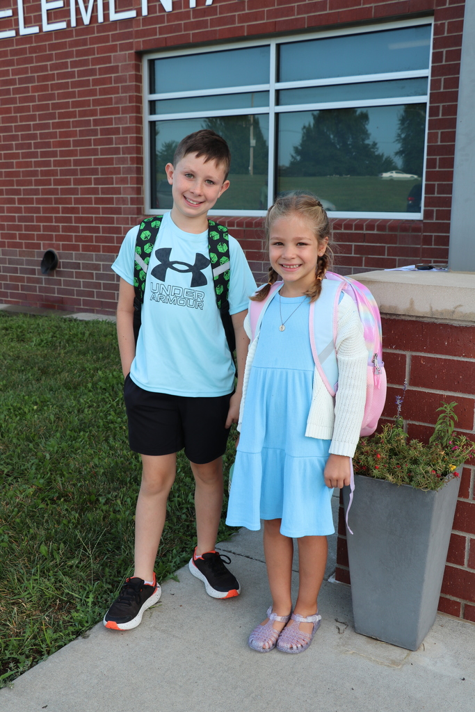 A boy and girl pose in front of the school with their backpacks on 