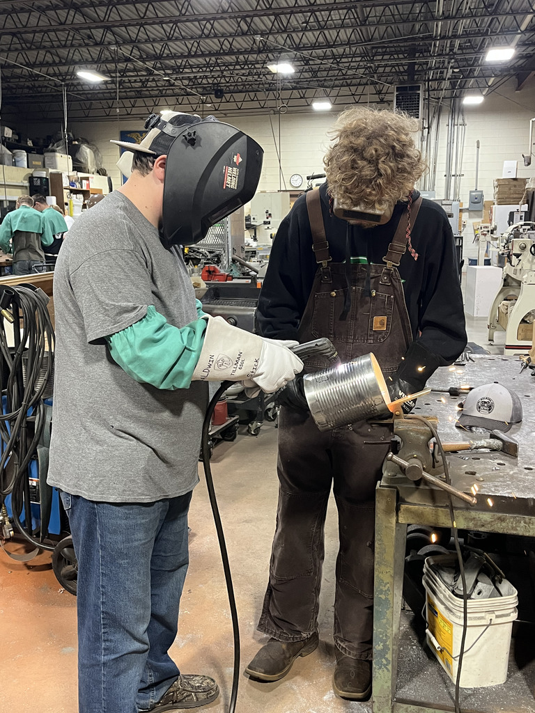 A student uses a heat tool to cut the face of his tin can jack-o-lantern. Another student watches.