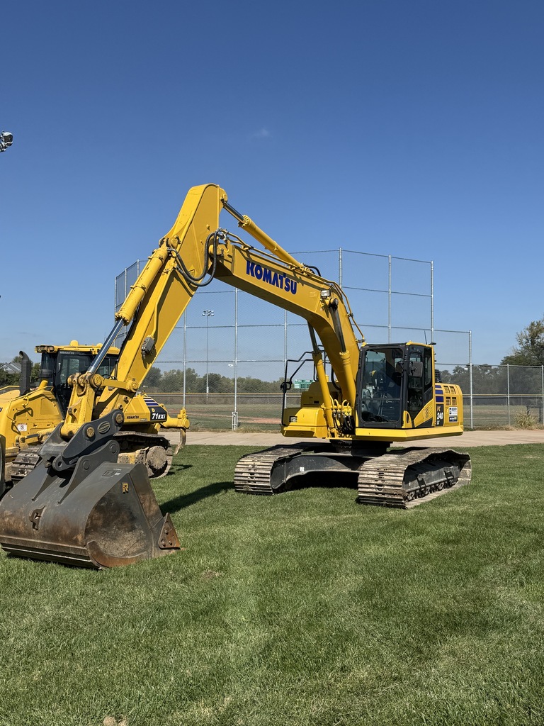 Large yellow Komatsu excavators are parked on the grass near a baseball field under a clear blue sky. The construction equipment is staged for the start of demolition and site work for the new Smithville Baseball and Softball Complex.