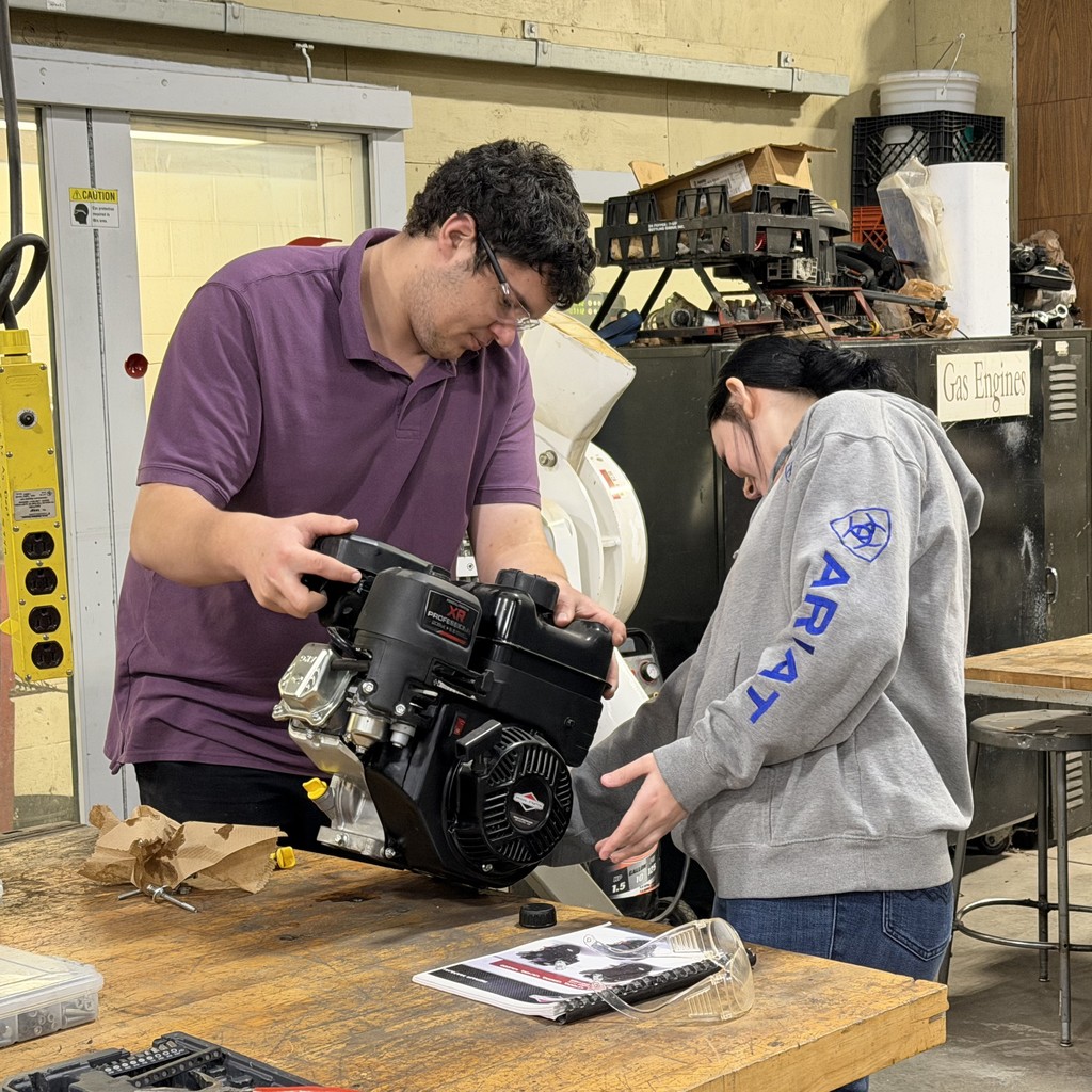 Two students stand at a workbench in a shop classroom, carefully handling a small engine as they prepare to take it apart and learn how it operates.