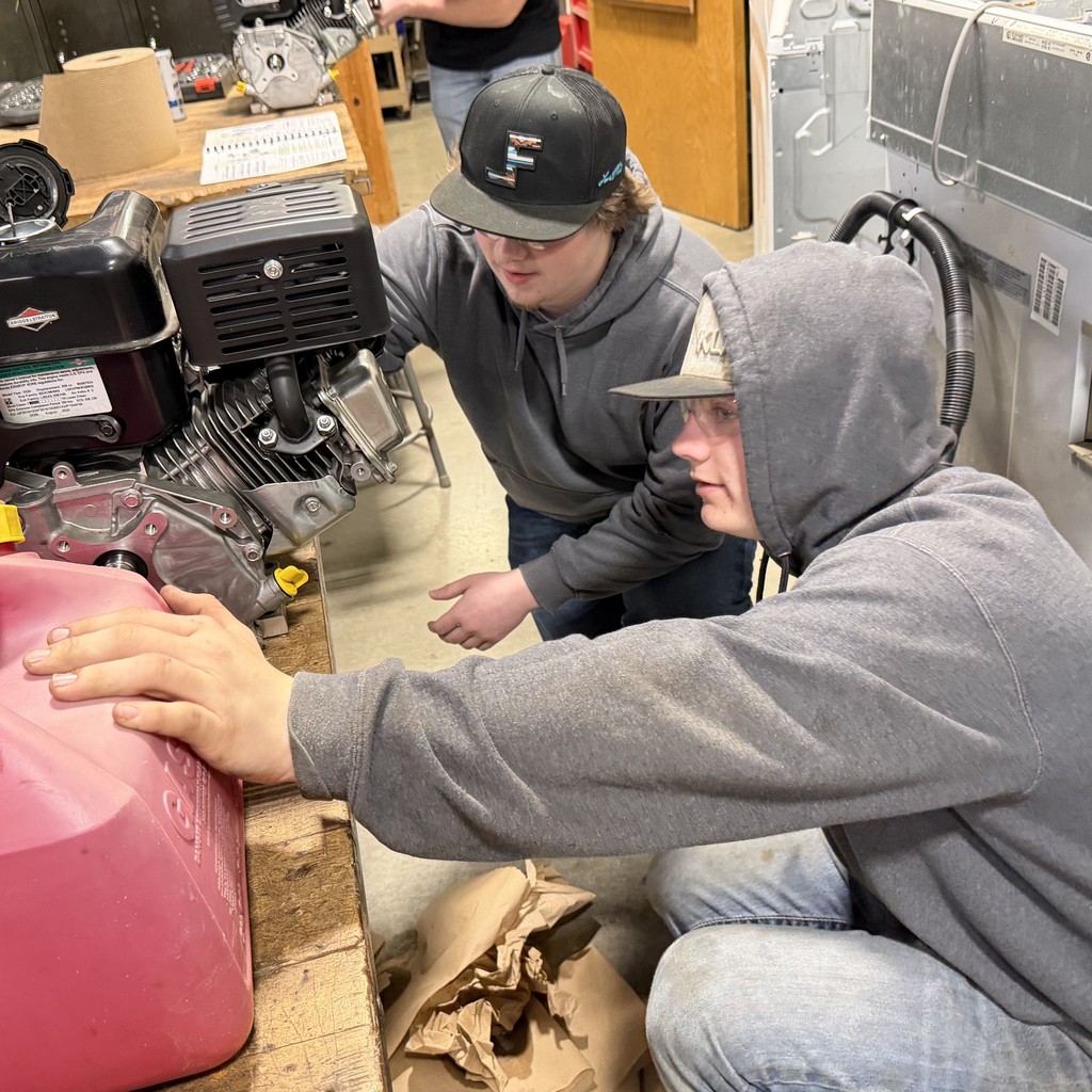 Two students work closely on a small engine in a shop setting, leaning in to inspect the engine housing and components during hands-on instruction.