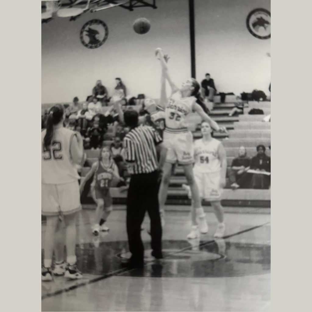 A referee and basketball players watch as two girls jump for the basketball.
