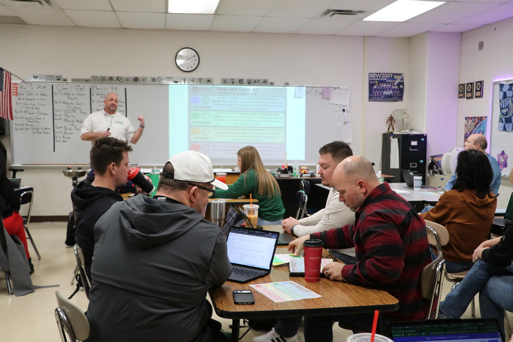 A teacher looks at a laptop. While another teacher speaks at the front of the classroom.