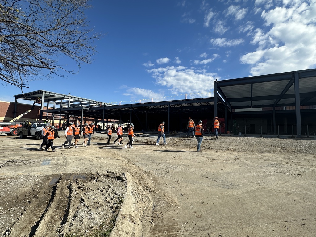 High School students dressed in orange vests and hard hats visit the construction site of the high school.