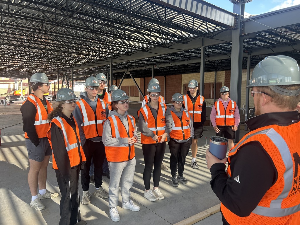 High School students dressed in orange vests and hard hats visit the construction site of the high school.