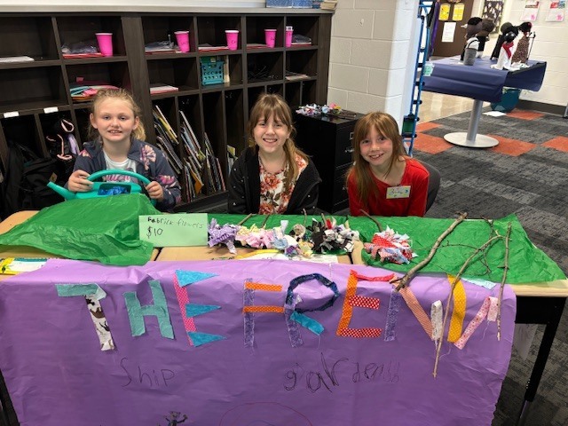 Three elementary school girls sit behind a table where they created a flower shop for their economics unit.