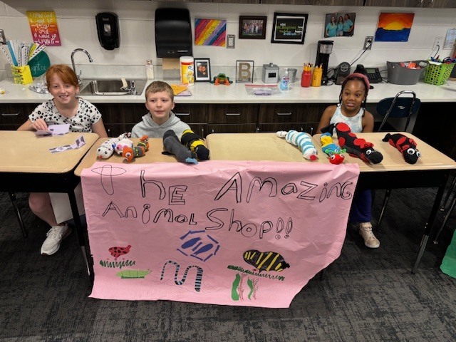 Three elementary school students sit behind a table where they created an animal shop for their economics unit.