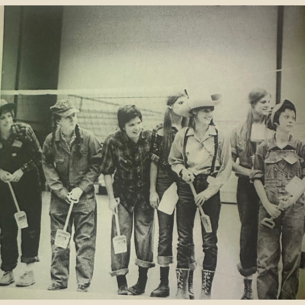 Black and white photo of a group of  students standing in a line, dressed in overalls, flannel shirts, and caps. Each person is holding a small shovel, participating in a group activity.