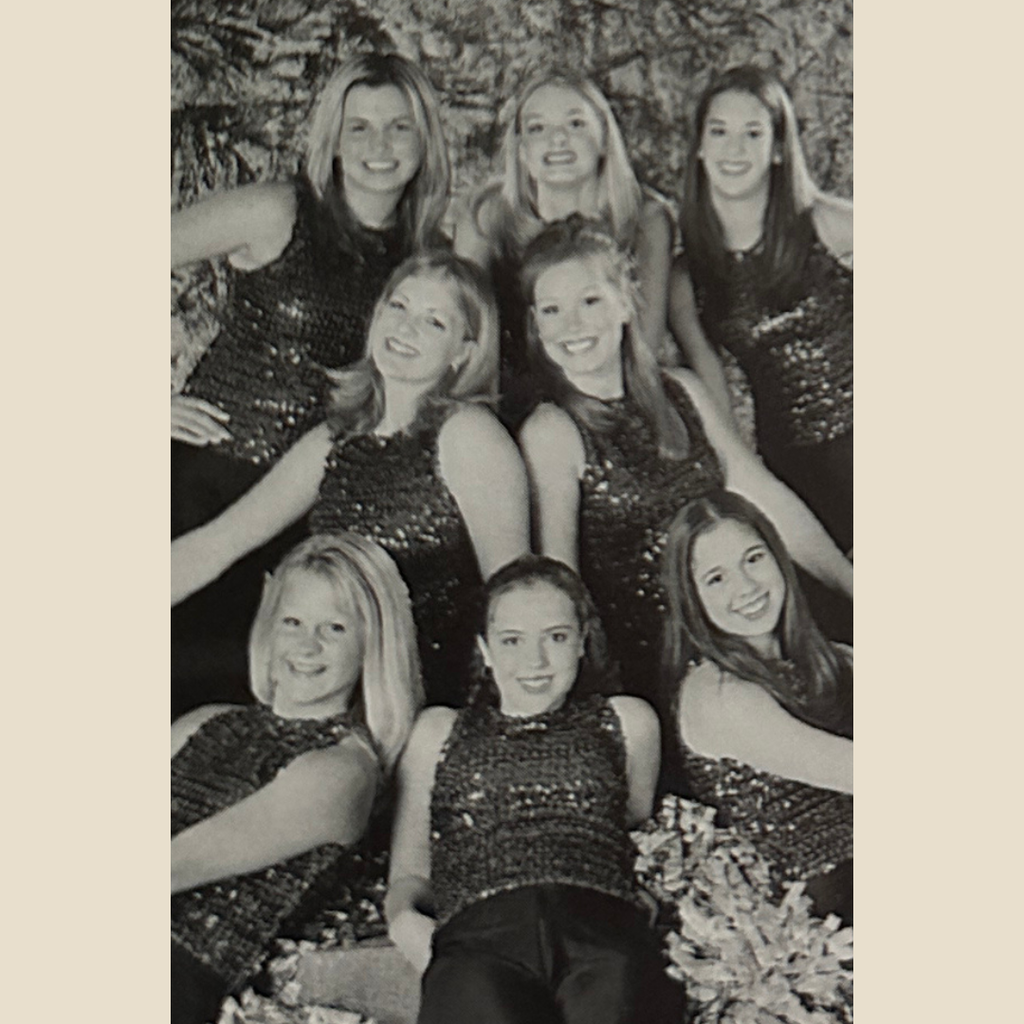 Group of 8 cheerleaders in glittery tops and pom poms in their hands all smile in a group for a picture