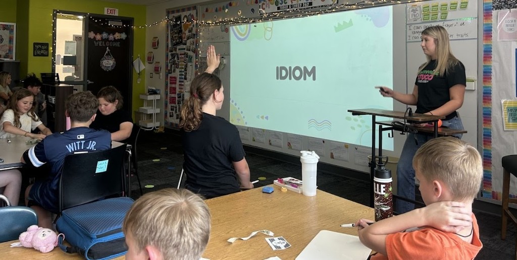 A high school student in the Cadet Teaching program at the front of the classroom during an ELA lesson with 5th graders. The word Idiom is on the board.