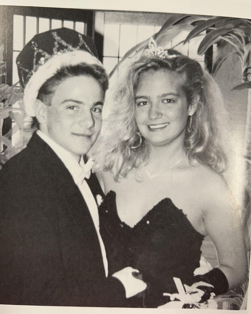 Black-and-white photo of a smiling young man and woman posing together indoors. The man wears a suit, bow tie, and a crown, while the woman wears a strapless dark dress, a tiara, and earrings. They stand close together in front of a window and leafy houseplants.