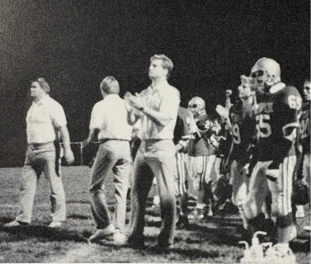 picture of coach Bill Maus clapping on the sideline with players behind him 